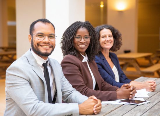 Happy diverse business team posing in street cafe. Business man and women with clasped hands sitting at table outside, looking at camera and smiling. Successful team concept